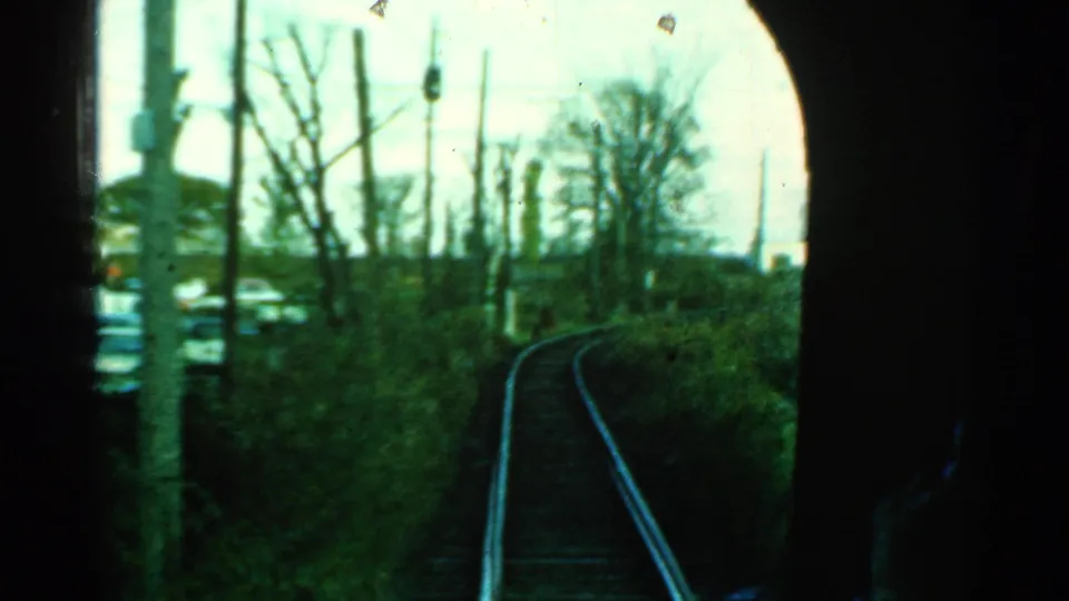 Dark-framed 8mm view from inside a train in Connecticut, with rails curving past brush, poles, bare trees, and a cluttered area.