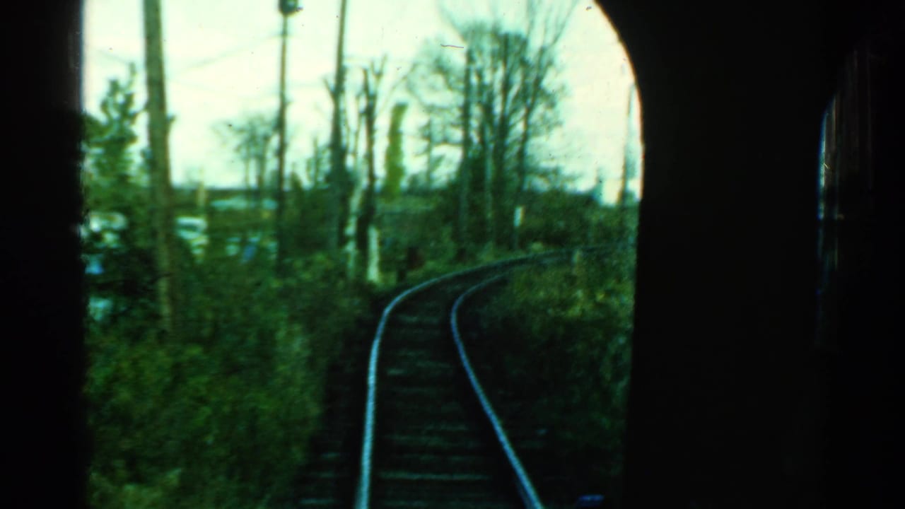 First-person 1953 Connecticut rail view with curved tracks, utility poles, bare trees, and green overgrowth beyond the train window.