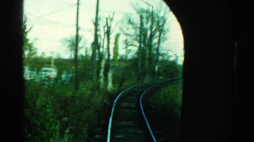 Vintage train-window view in Connecticut showing a curved rail line, dense roadside brush, bare trees, and cars in the background.