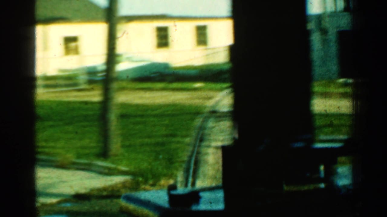 Passenger-window view in Connecticut, 1953, with dark interior framing around a light building, grass, sidewalk, and curving track.