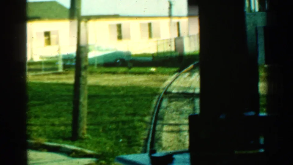 1953 Connecticut train-window frame with a light trackside building, utility pole, grass, sidewalk, and a parked car near the rail curve.
