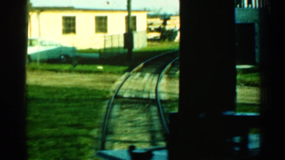 Connecticut 1953 passenger-view frame showing curved rails beside grass, dark train-window framing, a light building, and parked cars.