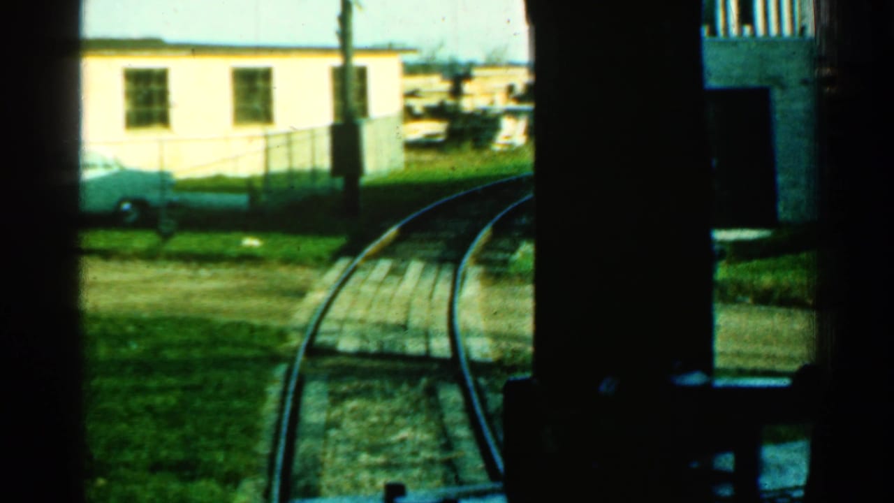 1953 Connecticut train POV through dark interior framing, showing curved rails, grass, a pale building, fencing, and a parked car.