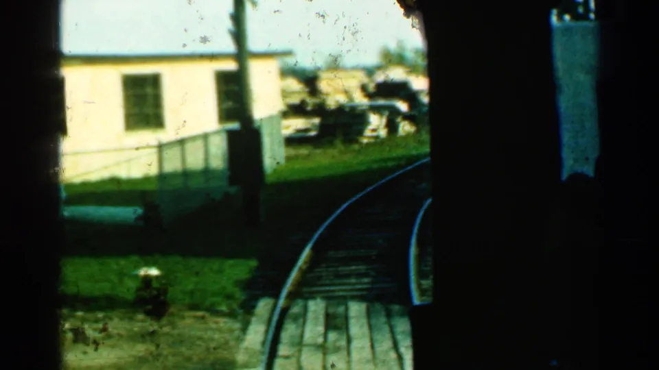 1953 Connecticut rail footage shows the train nearing a curve beside a pale building, chain-link fence, grass, and trackside materials.