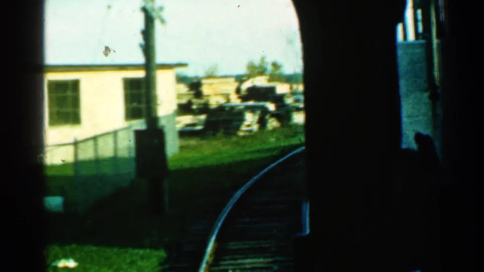 A late frame from the 1953 Connecticut train POV shows rails curving past a pale building, fence, grass, and scattered trackside objects.
