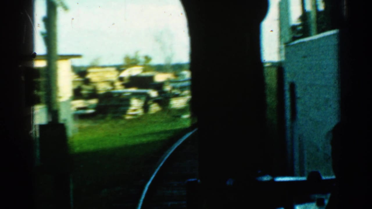 Train window view of a curve, poles, grass, a light building, and stacked trackside materials.