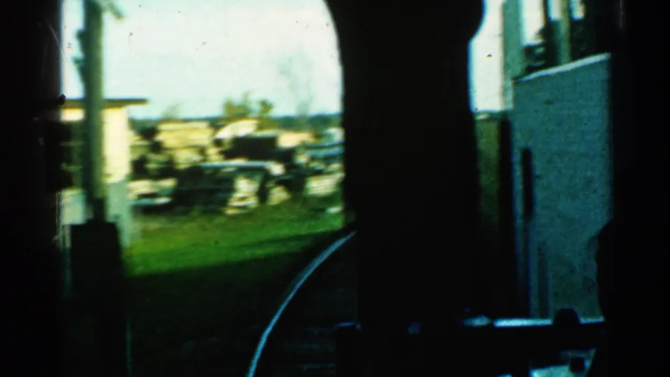From inside a moving train in Connecticut, the frame shows curved rail beside grass, utility poles, a light building, and stacked materials outside.
