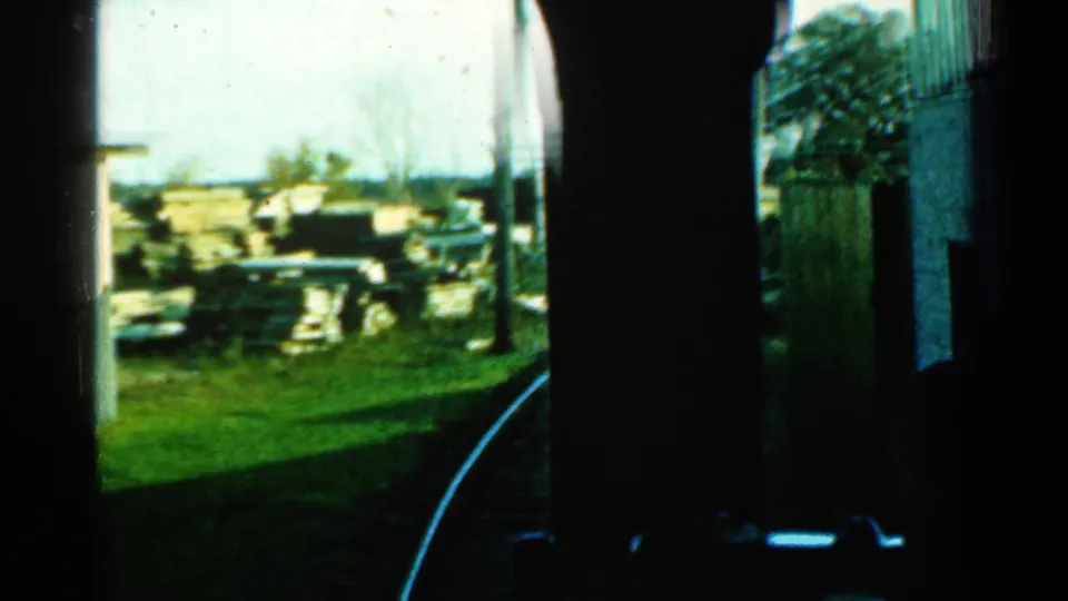 1953 Connecticut rail footage seen from a train window, with dark foreground framing, grass, a pole, stacked materials, and the track curving away.