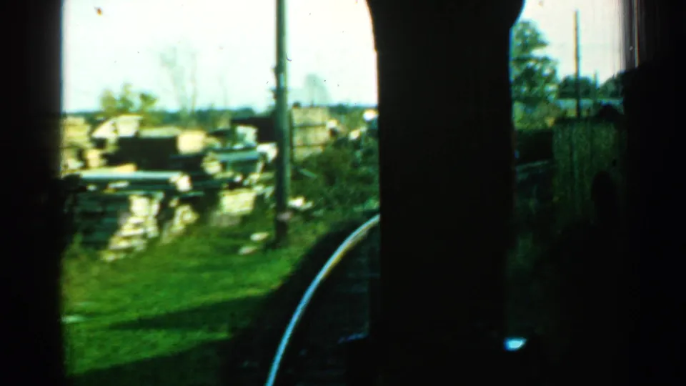 A Connecticut train-window view shows the rail curving along a grassy edge, with utility poles, stacked materials, and heavy shadow from the train interior.