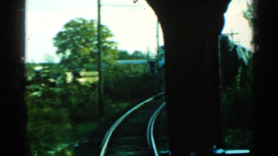 Final 1953 Connecticut window view of the rail bend, with trackside greenery, utility poles, and low structures beyond.