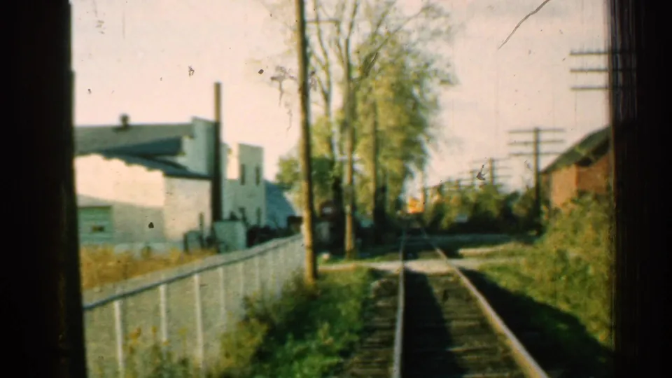 Moving train view in 1953 Connecticut, with rails leading past a white fence, trees, utility poles, and trackside buildings.