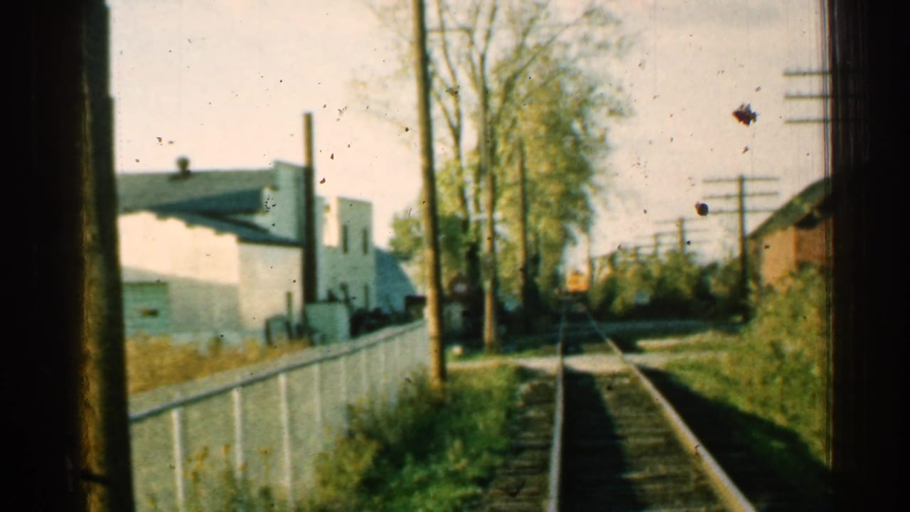 1953 Connecticut home-movie frame looking down railroad tracks from a train window, with a white fence, trees, utility poles, and trackside buildings.