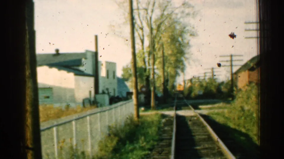 1953 Connecticut home-movie frame looking down railroad tracks from a train window, with a white fence, trees, utility poles, and trackside buildings.