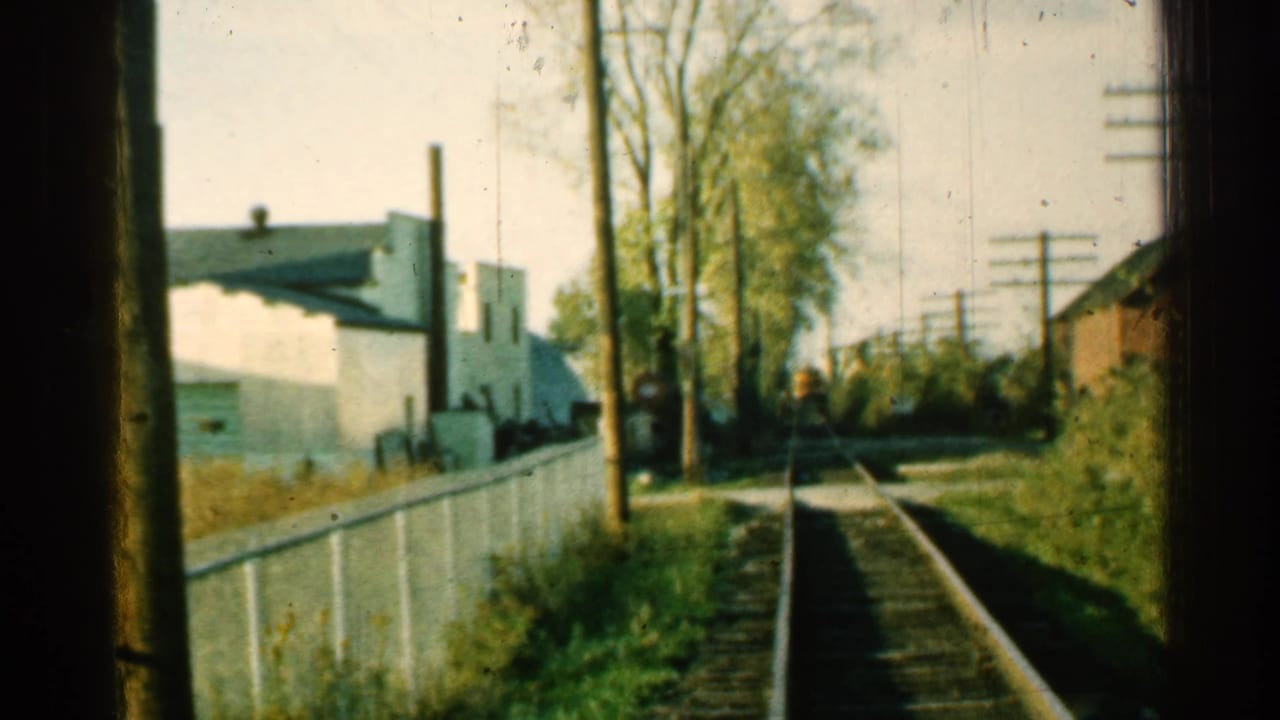 1953 Connecticut rail-side view from a moving train, showing track, white fence, trees, utility poles, and modest town buildings in soft 8mm color.