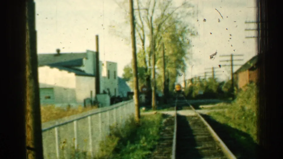 Final 1953 Connecticut home-movie frame of the railroad corridor, with track, white fence, trees, utility poles, grass, and small-town buildings.