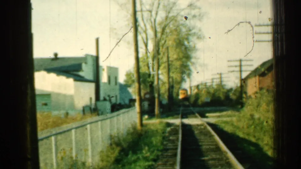 1953 Connecticut 8mm frame showing a distant train on tracks beside a white fence and low trackside buildings.
