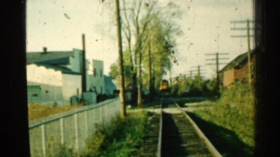 1953 Connecticut 8mm frame of a train still far along the tracks in a quiet suburban rail-side setting.
