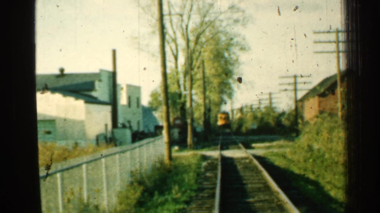 Yellow train seen ahead on tracks beside a white fence, utility poles, trees, and low buildings.