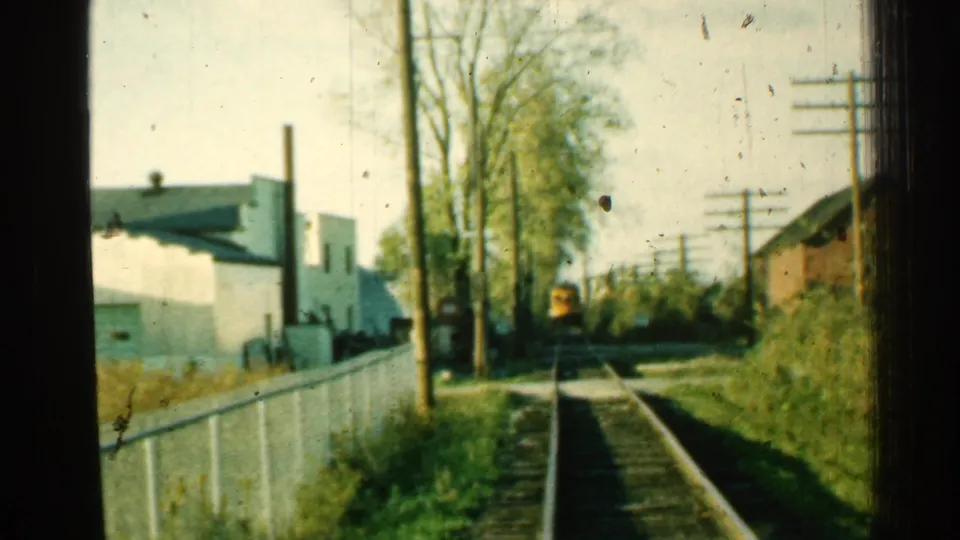 1953 Connecticut 8mm frame showing a yellow train ahead on rural tracks with fencing, trees, utility poles, and low buildings.