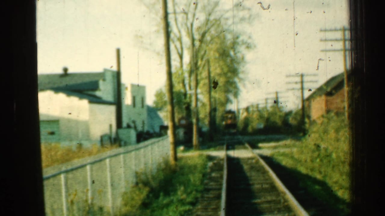 1953 Connecticut 8mm frame with tracks leading toward a yellow train, bordered by a white fence, trees, poles, and small buildings.