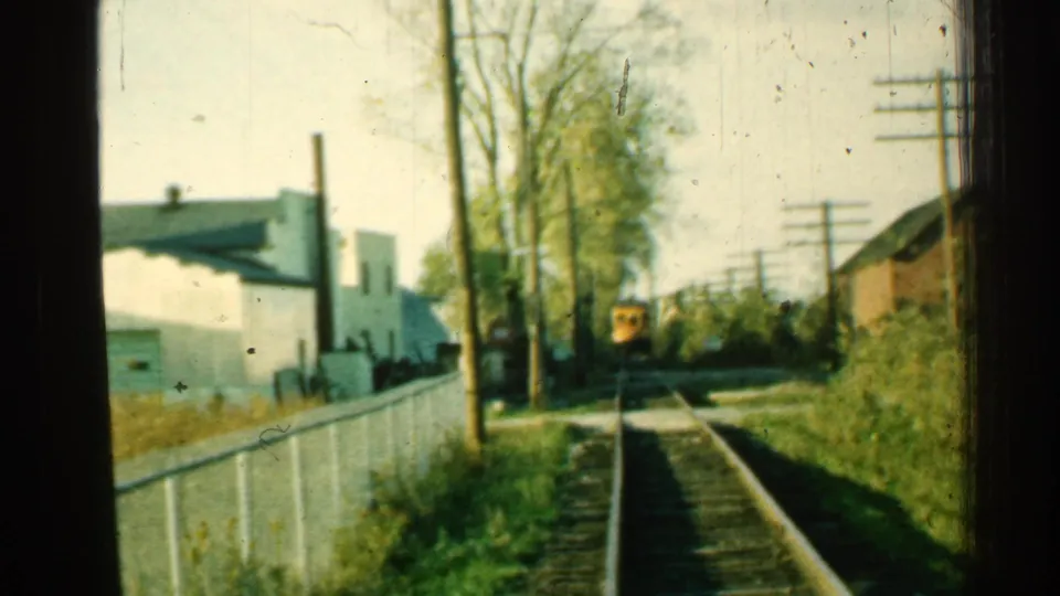 1953 Connecticut 8mm frame showing a distant yellow train along tracks bordered by fencing, trees, utility poles, and low buildings.