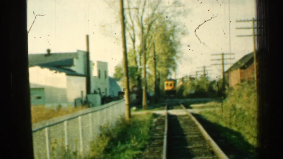 1953 Connecticut 8mm frame of a yellow train farther down a rural rail line with fence, trees, utility poles, and buildings.