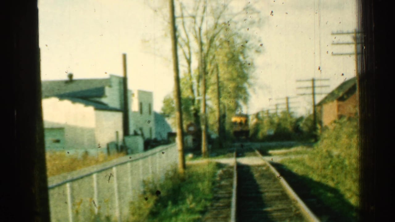 Railroad tracks run past a fence and white building toward a small yellow train car in the distance.