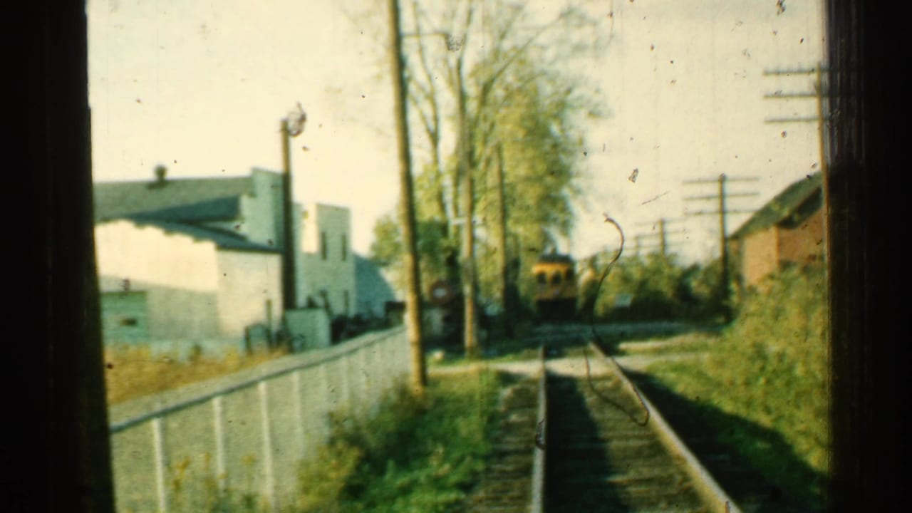 Soft 1953 Connecticut home-movie frame with railroad tracks, utility poles, trees, and a yellow train car beyond a fenced trackside area.
