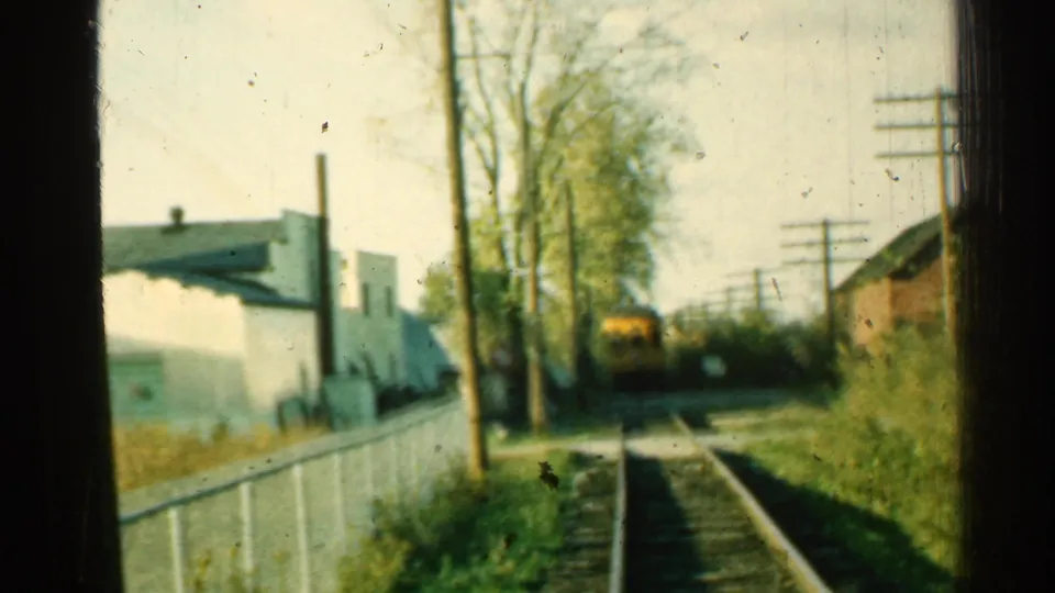 1953 Connecticut 8mm view of a rural or industrial track corridor with rails, fencing, utility poles, trees, and a yellow train car ahead.
