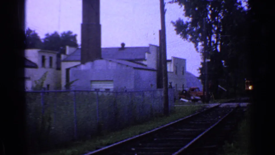 Darker 1953 Connecticut 8mm frame showing railroad tracks beside fencing, a white industrial building, a tall chimney, a utility pole, and trees.
