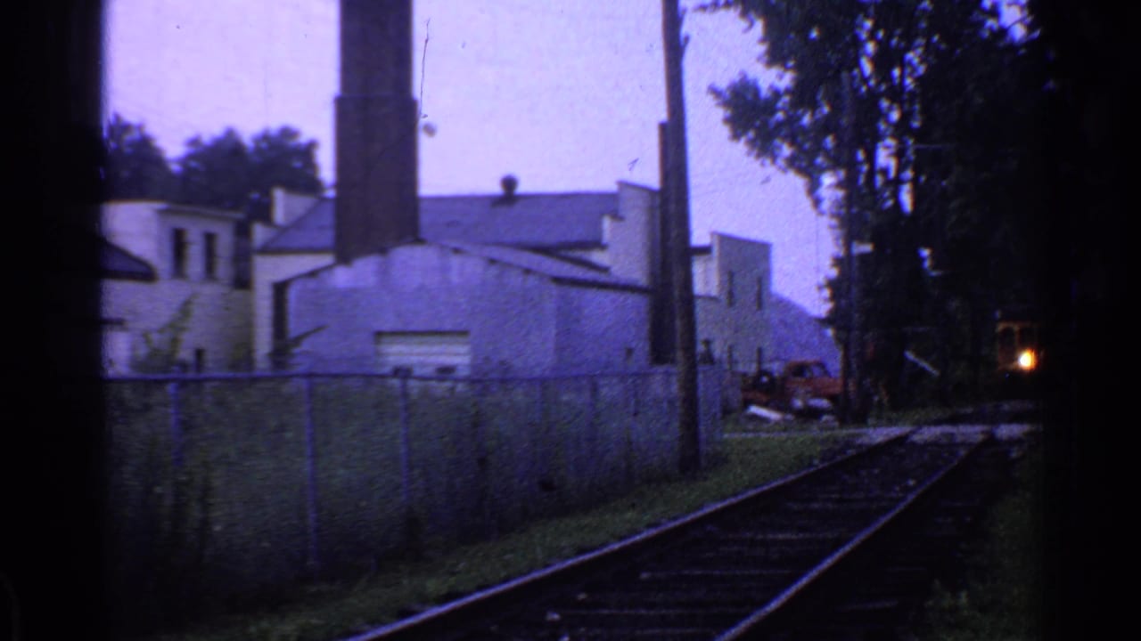 1953 Connecticut 8mm frame showing rail tracks beside fencing, trees, utility poles, and an industrial building with a tall chimney.