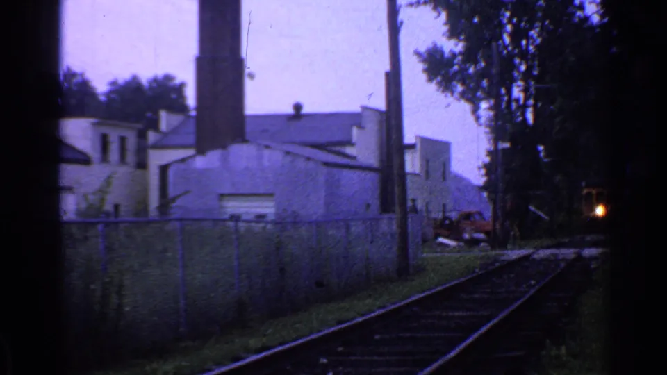 1953 Connecticut 8mm frame showing rail tracks beside fencing, trees, utility poles, and an industrial building with a tall chimney.