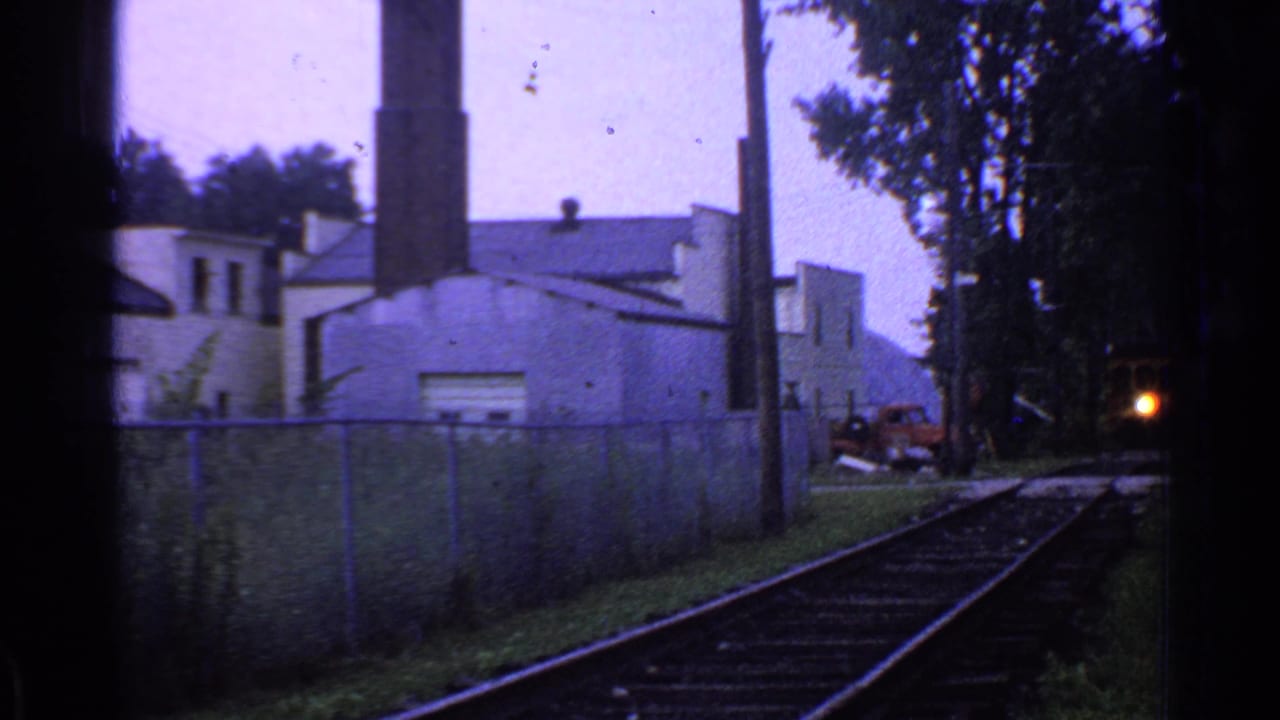 A small bright train headlight appears in the distance on Connecticut rail tracks beside a chain-link fence and industrial building.