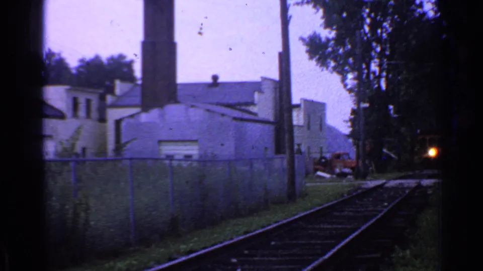 A small bright train headlight appears in the distance on Connecticut rail tracks beside a chain-link fence and industrial building.