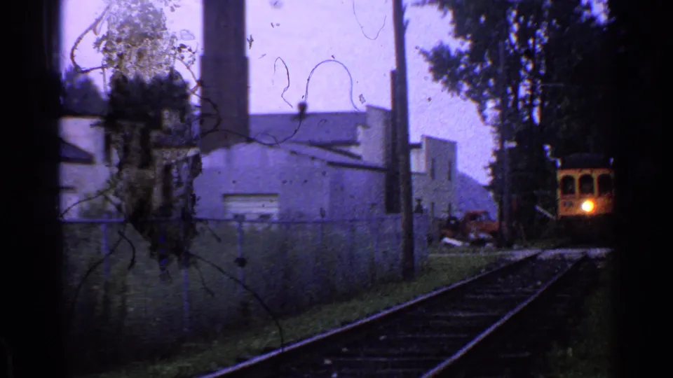 The yellow train nears the camera on tracks beside a Connecticut industrial building, with a bright headlight and red vehicle nearby.