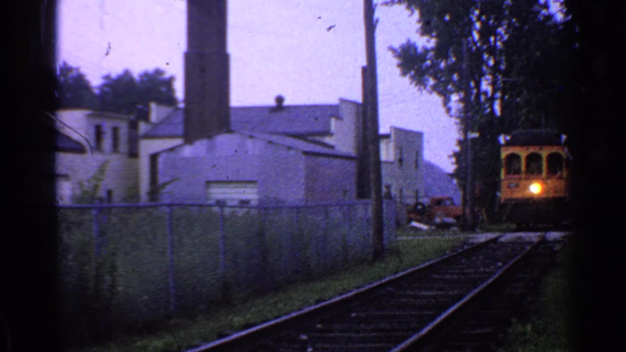 1953 Connecticut 8mm frame showing a yellow train or trolley approaching along tracks past a fence, smokestack, and low industrial buildings.