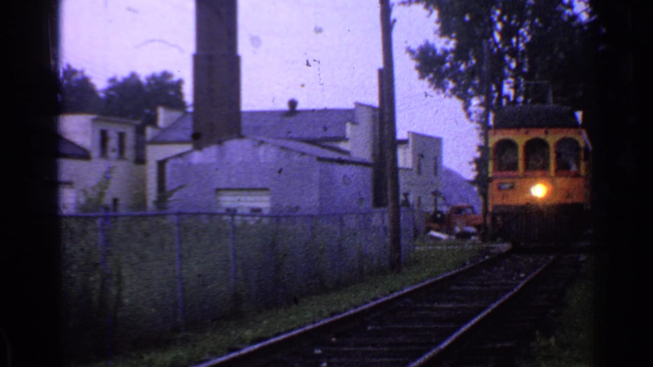 1953 Connecticut frame of a yellow train or trolley moving closer on rails beside a fence, with a lit headlight and smokestack in the background.