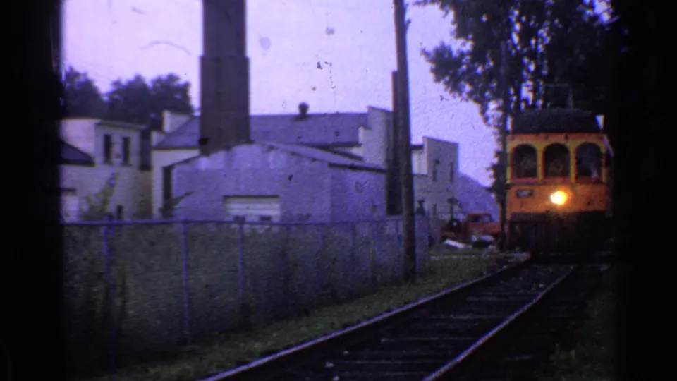 1953 Connecticut frame of a yellow train or trolley moving closer on rails beside a fence, with a lit headlight and smokestack in the background.