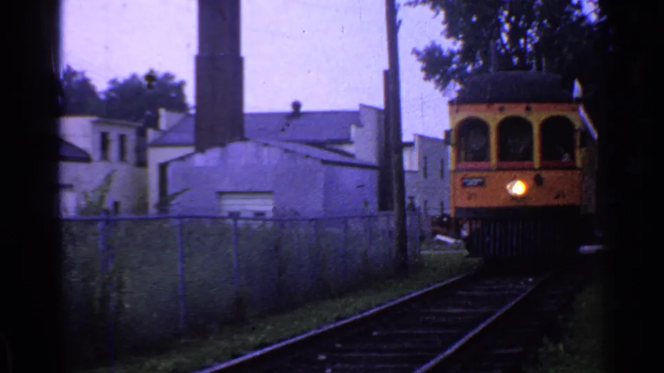 1953 Connecticut frame showing the yellow rail vehicle partly at right as it passes along tracks beside industrial buildings and a chain-link fence.
