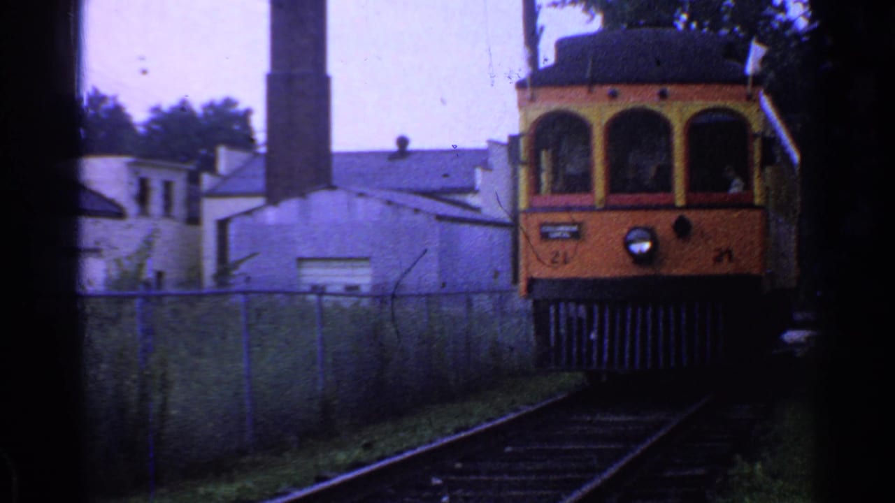 Orange and yellow trolley in Connecticut moves closer to the camera, with rails, fence, buildings, and a smokestack visible behind it.