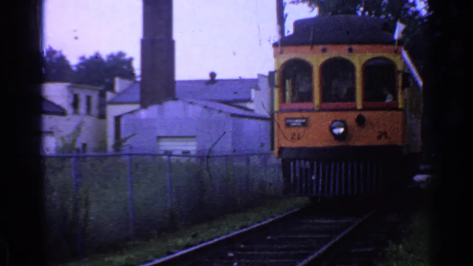 Orange and yellow trolley in Connecticut moves closer to the camera, with rails, fence, buildings, and a smokestack visible behind it.