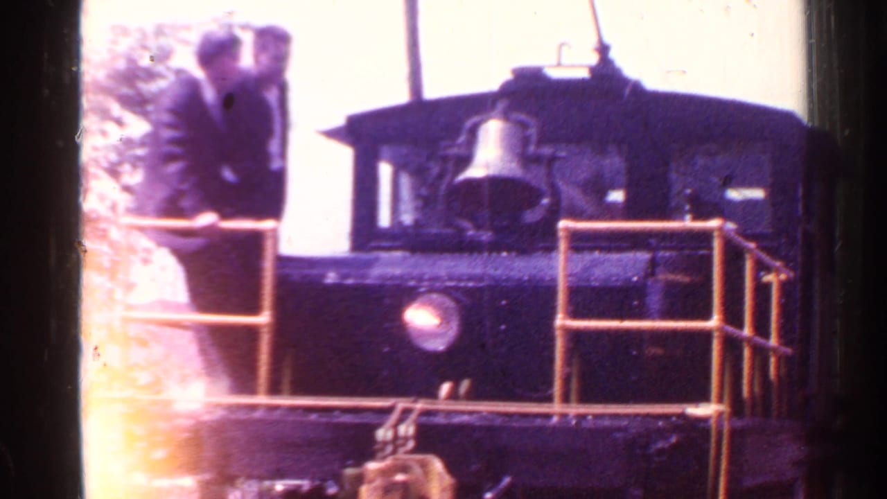 Man in a dark suit stands on the front platform of a small locomotive with railings.