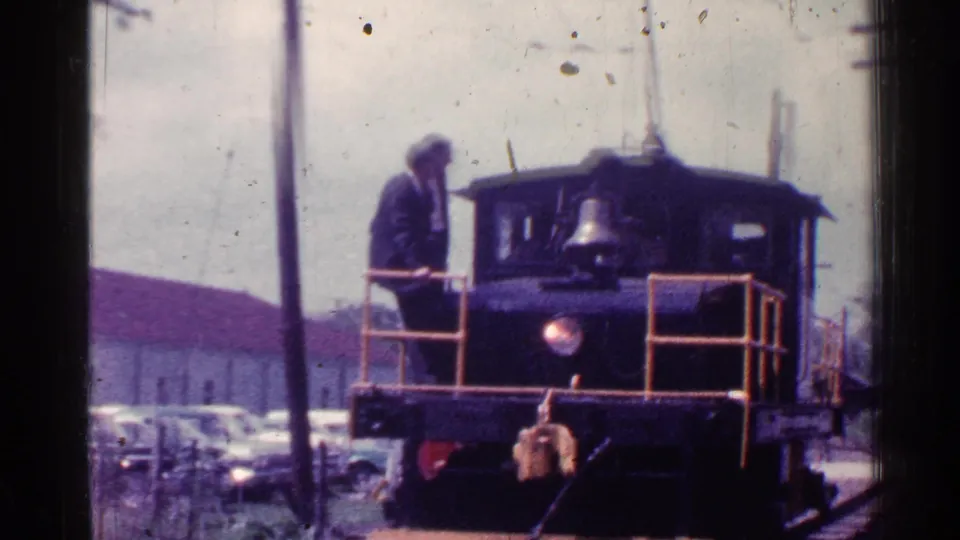 1953 Connecticut 8mm frame showing a small locomotive, suited man, parked cars, utility poles, and a red-roofed building.