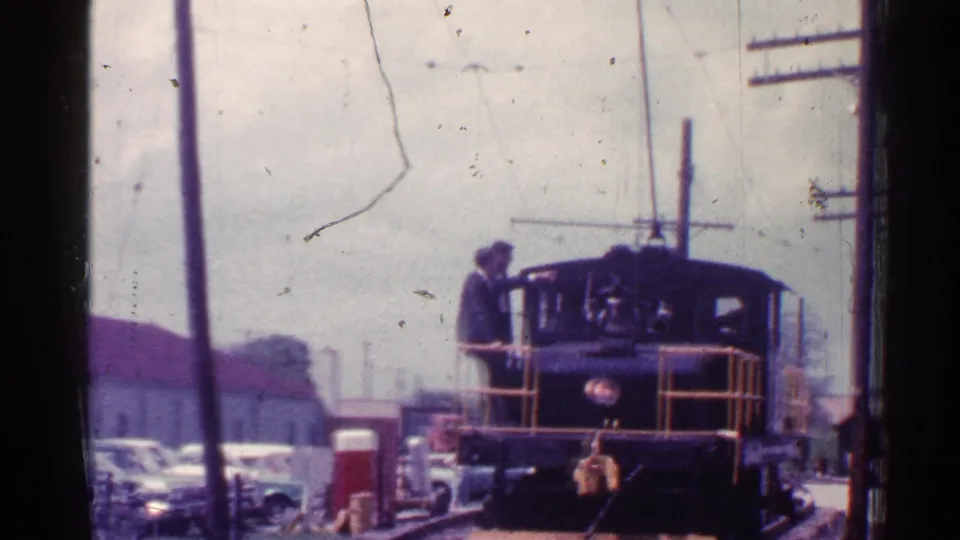 1953 Connecticut 8mm frame with a wider view of a small locomotive at tracks, a suited man, parked cars, and utility poles.