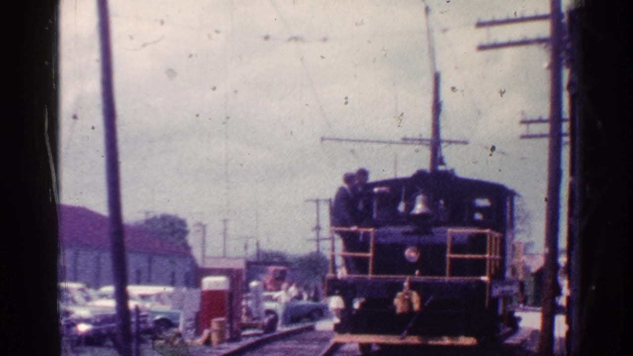Small railcar on tracks with a person standing near the cab, parked cars, and utility poles nearby.