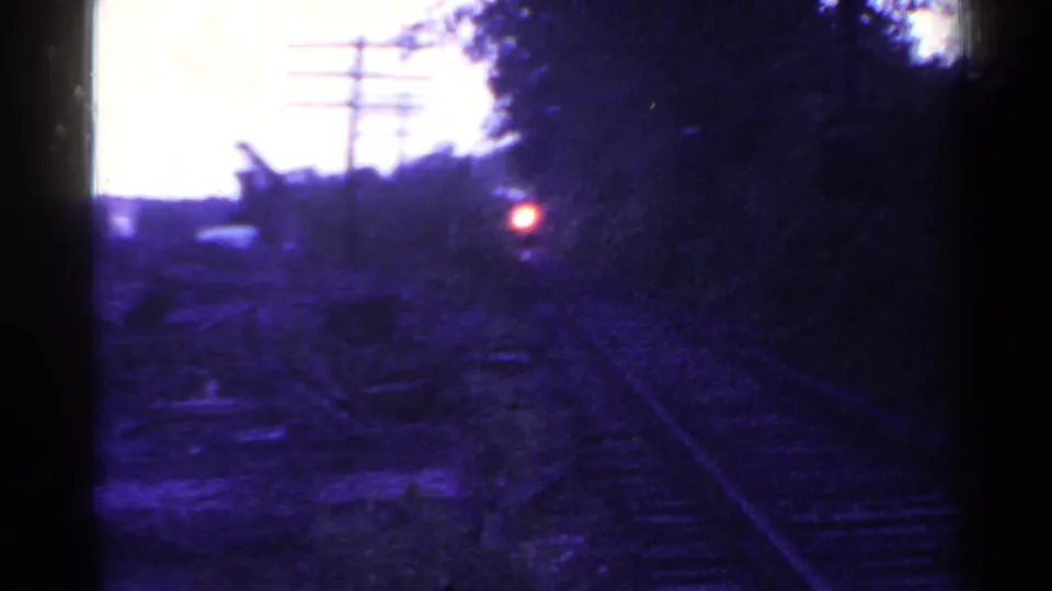 Shadowed home-movie frame of Connecticut rail tracks, utility poles, trees, and a small red light farther down the line.
