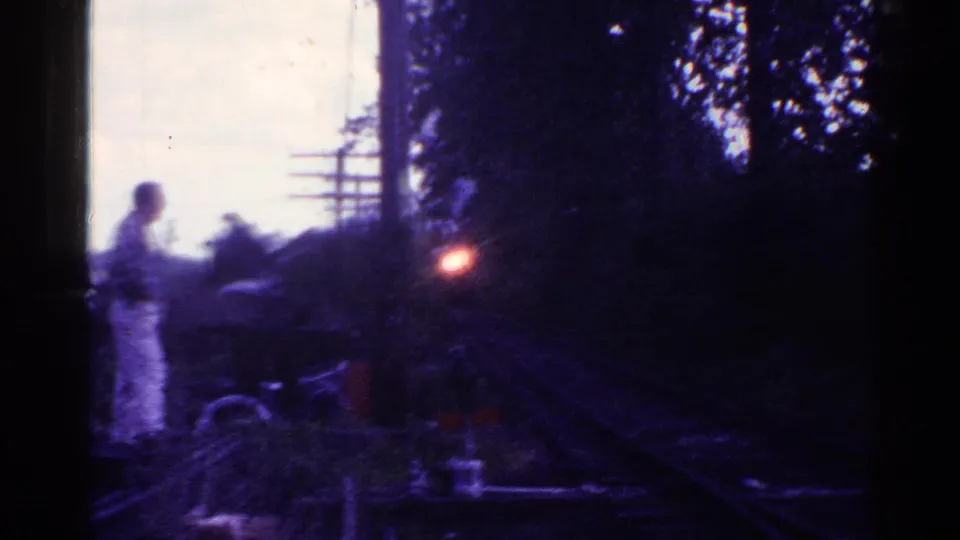 A man in light clothing stands beside Connecticut railroad tracks as a train light approaches in 1953 home-movie footage.