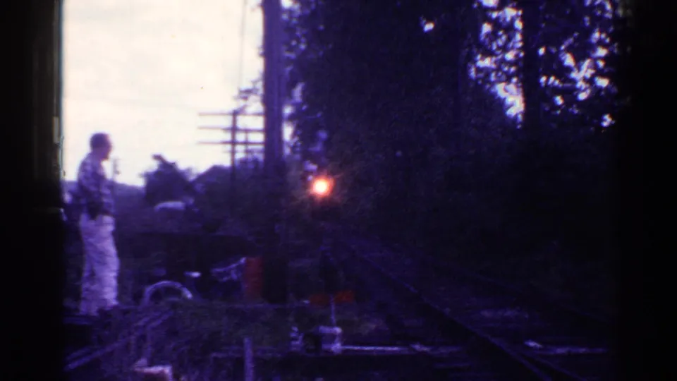 1953 Connecticut frame showing a man near railroad tracks, a bicycle, utility poles, trees, and the red light of an approaching train.