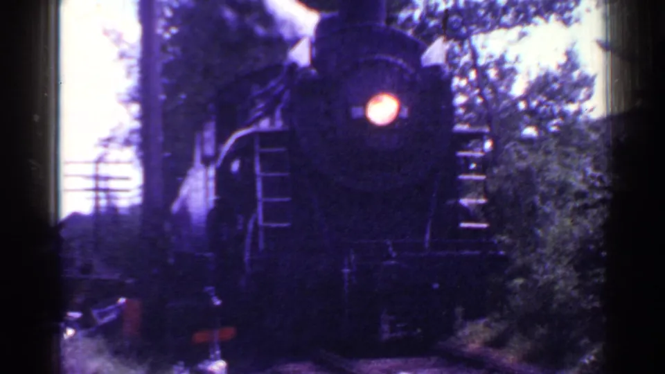 1953 Connecticut 8mm footage shows a steam locomotive approaching through trees, its headlight bright against the dark, purple-shifted film.