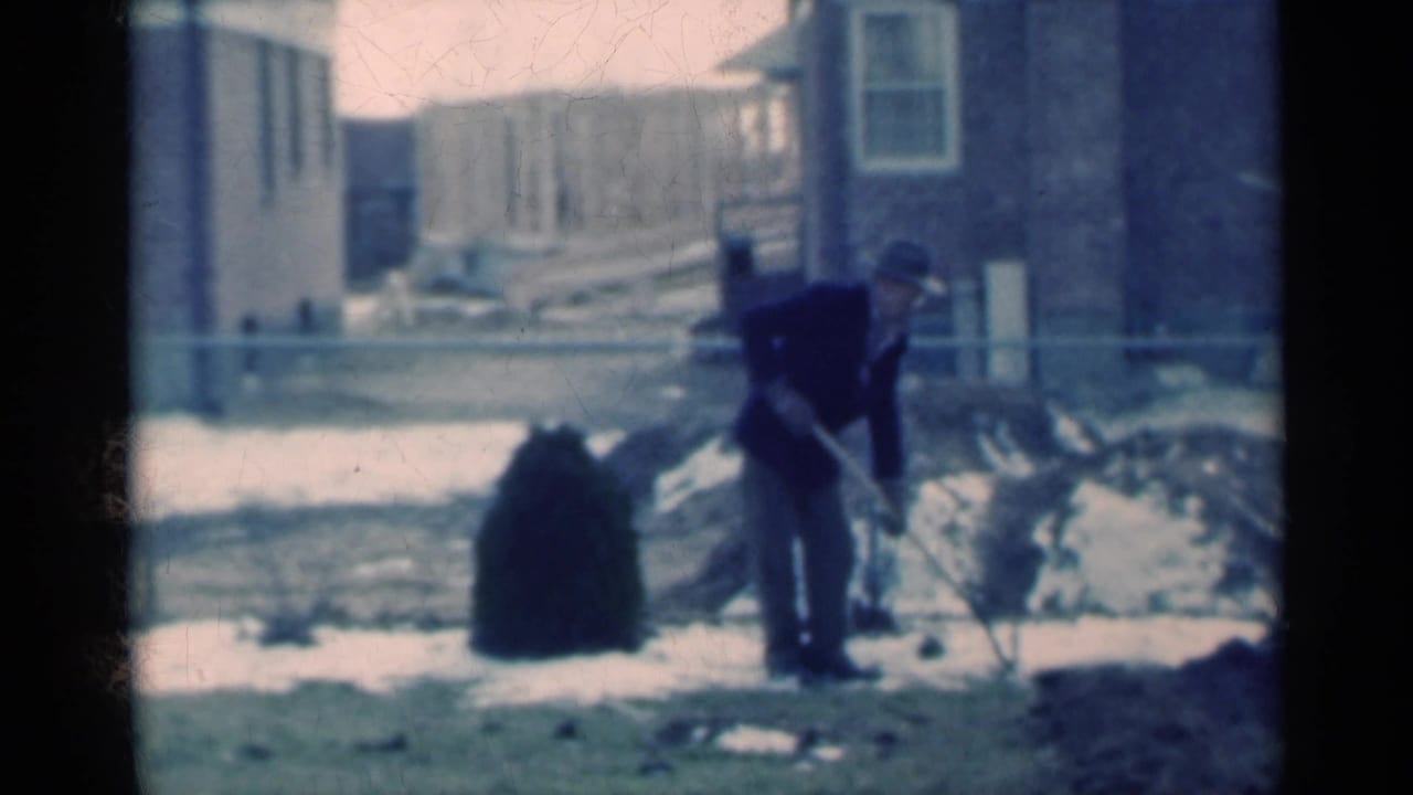 In 1939 Michigan home-movie footage, a man in a dark coat and hat shovels dirt beside patchy snow in a yard.